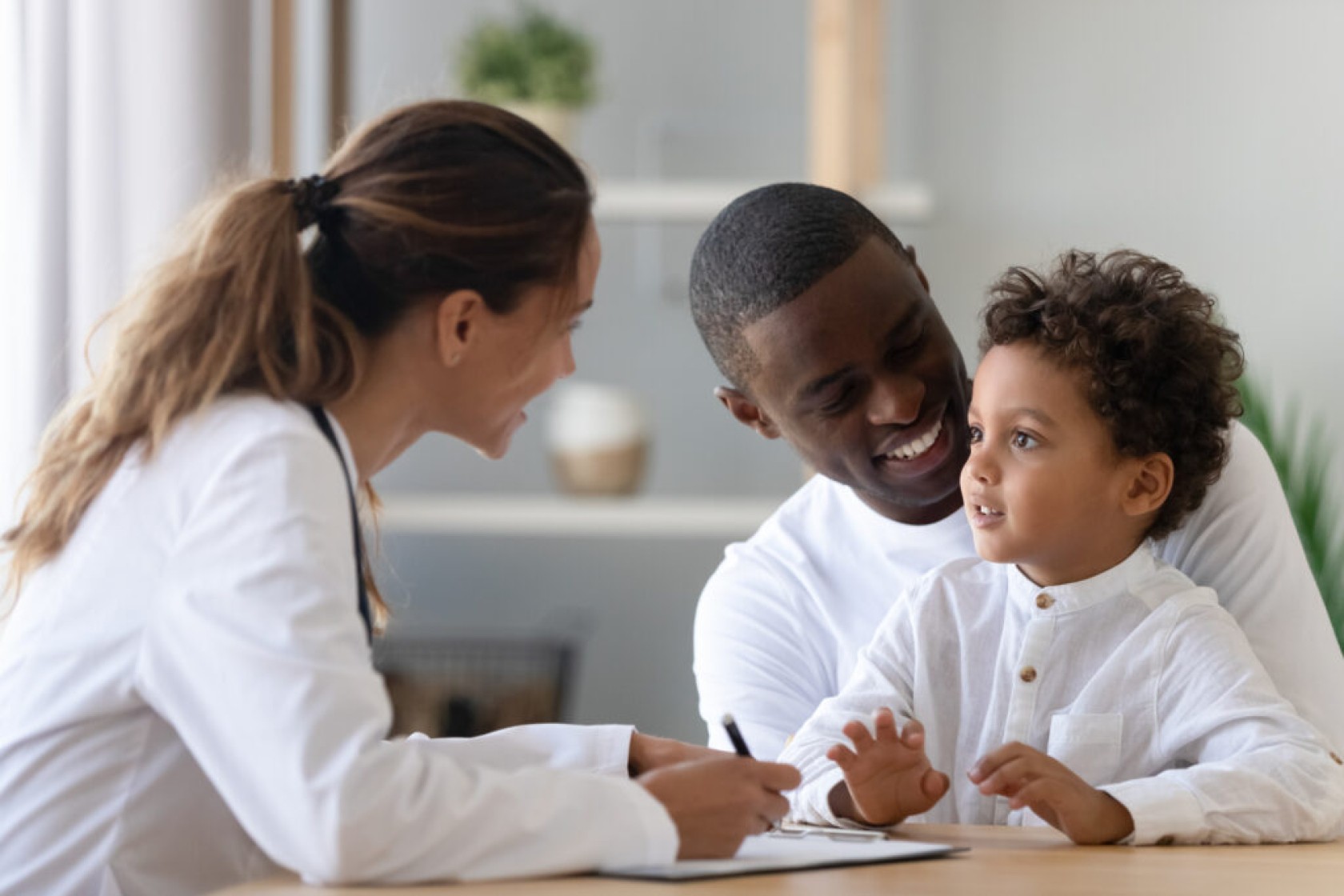 Cheerful young female pediatrician and african american smiling father listening to mixed race little patient, telling doctor about well-being. Happy multiracial family visiting clinic for check up.